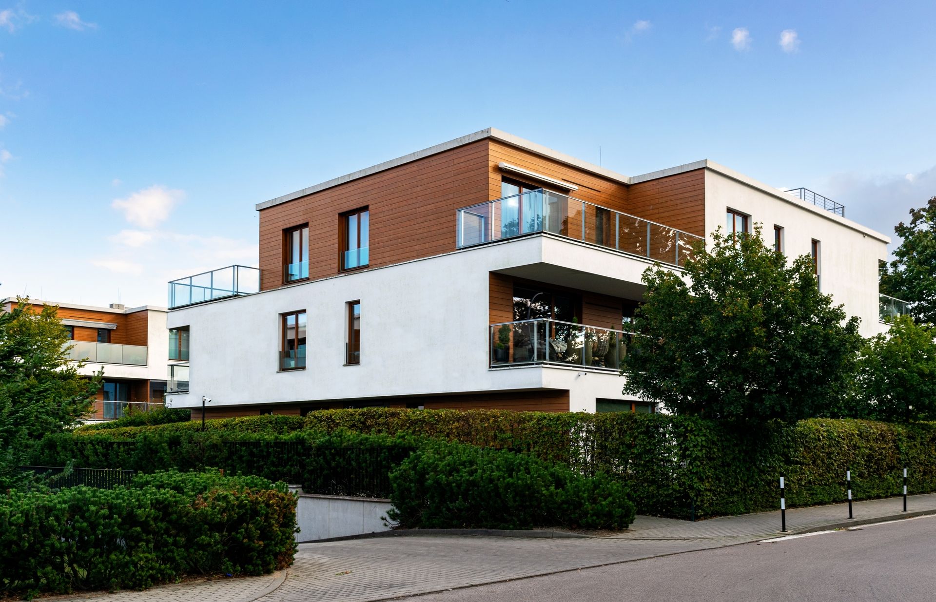 Modernes Mehrfamilienhaus mit weißer Fassade, Holzverkleidung und Glasbalkonen, umgeben von gepflegten Hecken und Bäumen